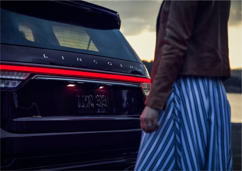 A person is shown near the rear of a 2023 Lincoln Aviator® SUV as the Lincoln Embrace illuminates the rear lights | Stivers Lincoln in Waukee IA