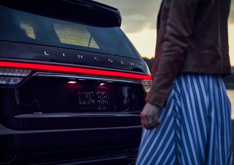 A person is shown near the rear of a 2024 Lincoln Aviator® SUV as the Lincoln Embrace illuminates the rear lights | Stivers Lincoln in Waukee IA